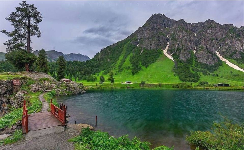 A serene lake surrounded by lush green hills and pine trees in Astore Valley, Gilgit-Baltistan, with a small wooden bridge extending over the water and dramatic mountains in the background.Cold Places in Pakistan