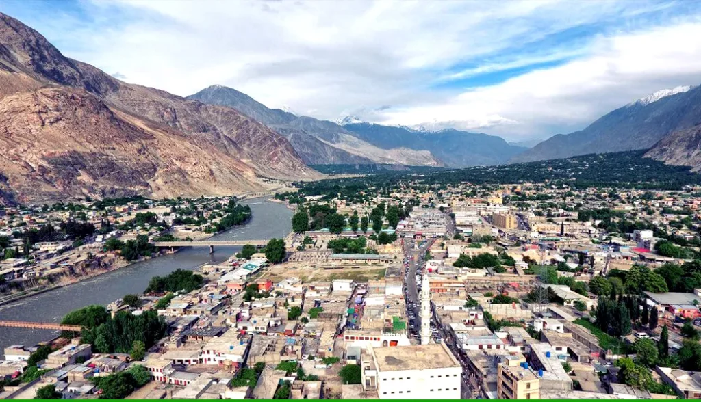 Aerial view of Gilgit City surrounded by rugged mountains, with the Gilgit River flowing through the valley and a mix of urban buildings, roads, and green patches under a partly cloudy sky.