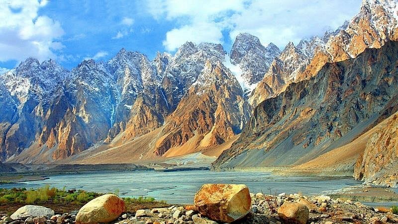 A panoramic view of Hunza Valley featuring sharp, rocky mountain peaks under a bright blue sky, with a calm river flowing through the valley and scattered boulders in the foreground.
