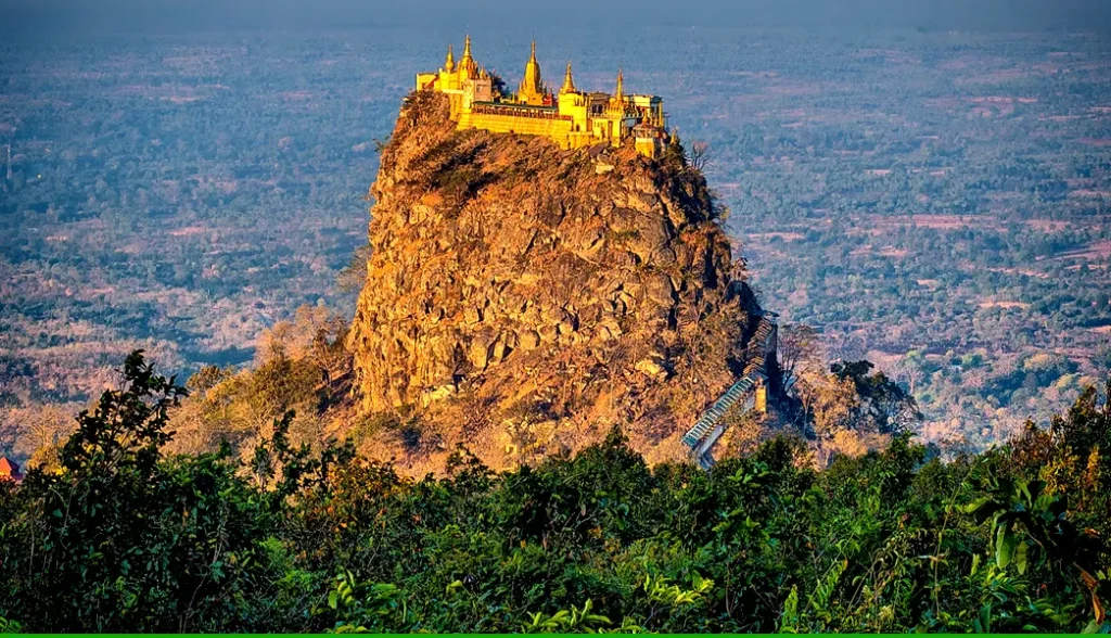 A dramatic rocky hilltop in Kalat, Balochistan, glowing in golden sunlight with a historic structure perched atop, surrounded by dense green foliage and expansive valley views in the background.