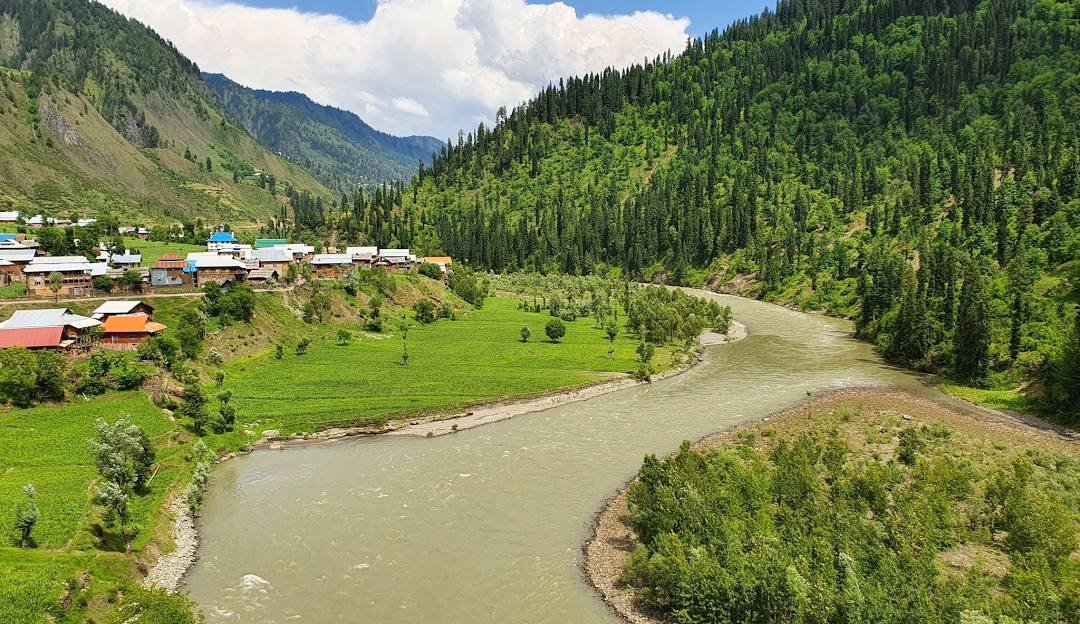 A scenic view of a curving river flowing through a lush green valley in Azad Kashmir, Pakistan. Traditional houses with colorful rooftops are nestled against forested hillsides, surrounded by pine-covered mountains under a bright sky with clouds.