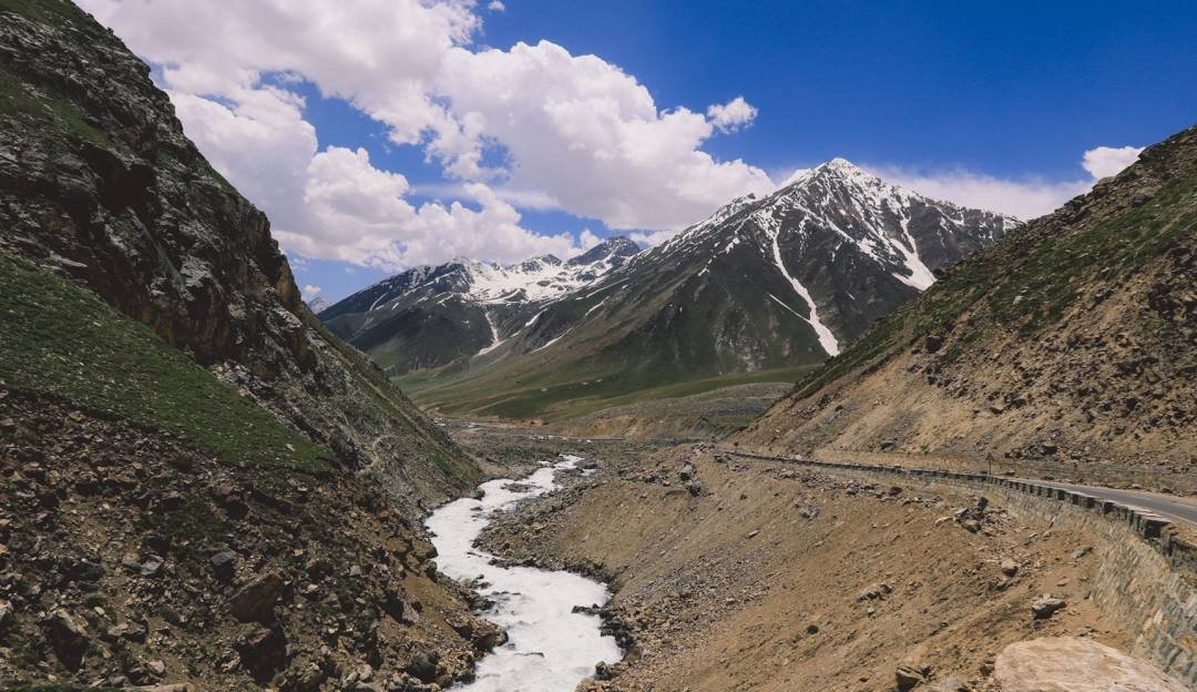 A breathtaking view of a snow-capped mountain range in Gilgit-Baltistan, Pakistan, with a winding river cutting through the rocky valley below and a paved road running alongside the hillside under a bright blue sky with scattered clouds.
