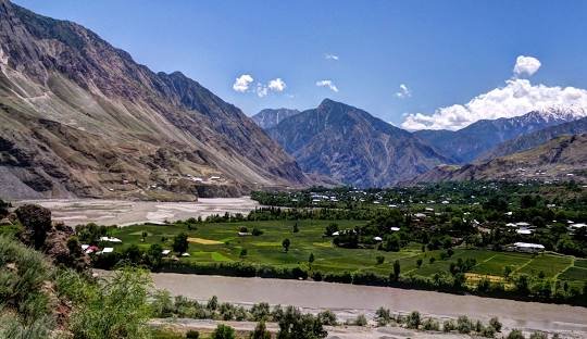 A panoramic view of a fertile green valley in Khyber Pakhtunkhwa, Pakistan, surrounded by majestic mountains. The image shows a river flowing through the valley, agricultural fields, and scattered houses nestled in the landscape under a clear blue sky with scattered clouds.