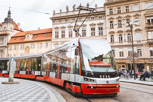 Red and white modern tram navigating the cobbled streets of Old Town Prague, with historic buildings and pedestrians in the background, representing the efficiency of Czech public transportation.