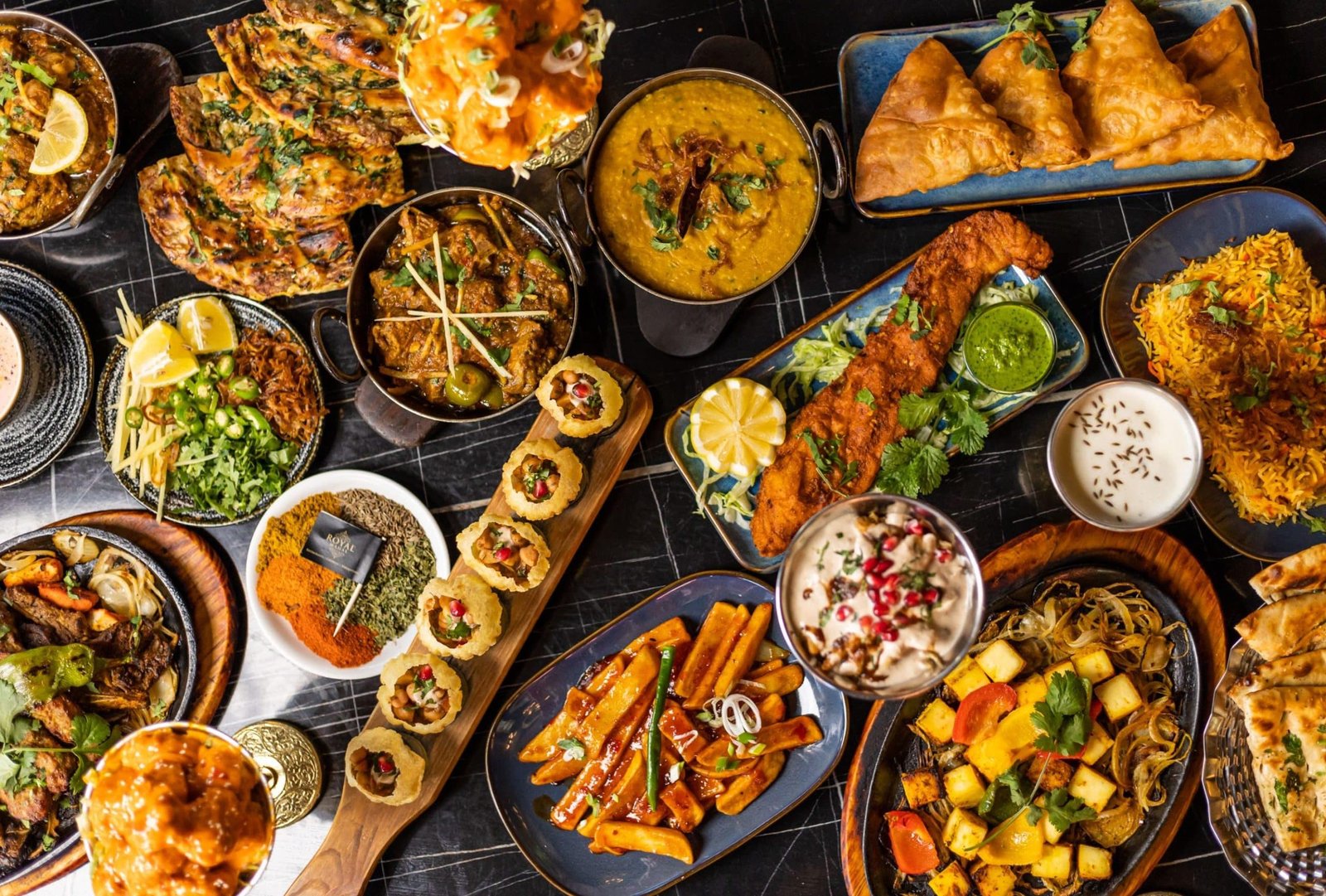 Top view of a vibrant assortment of Pakistani dishes including curries, grilled meats, rice, samosas, and desserts, artistically arranged on a dark table setting