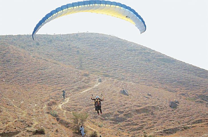 A person paraglides over the dry, rugged hills near Khanpur Dam, Pakistan, with a yellow and blue parachute canopy and a few people watching from the ground below. Paragliding in Khanpur Dam