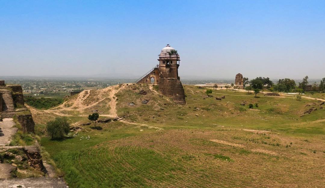 Historic watchtower of Rohtas Fort, perched on a hill overlooking the landscape of Punjab, Pakistan. The image showcases the fort's ancient architecture, surrounding open fields, and distant ruins under a bright blue sky.