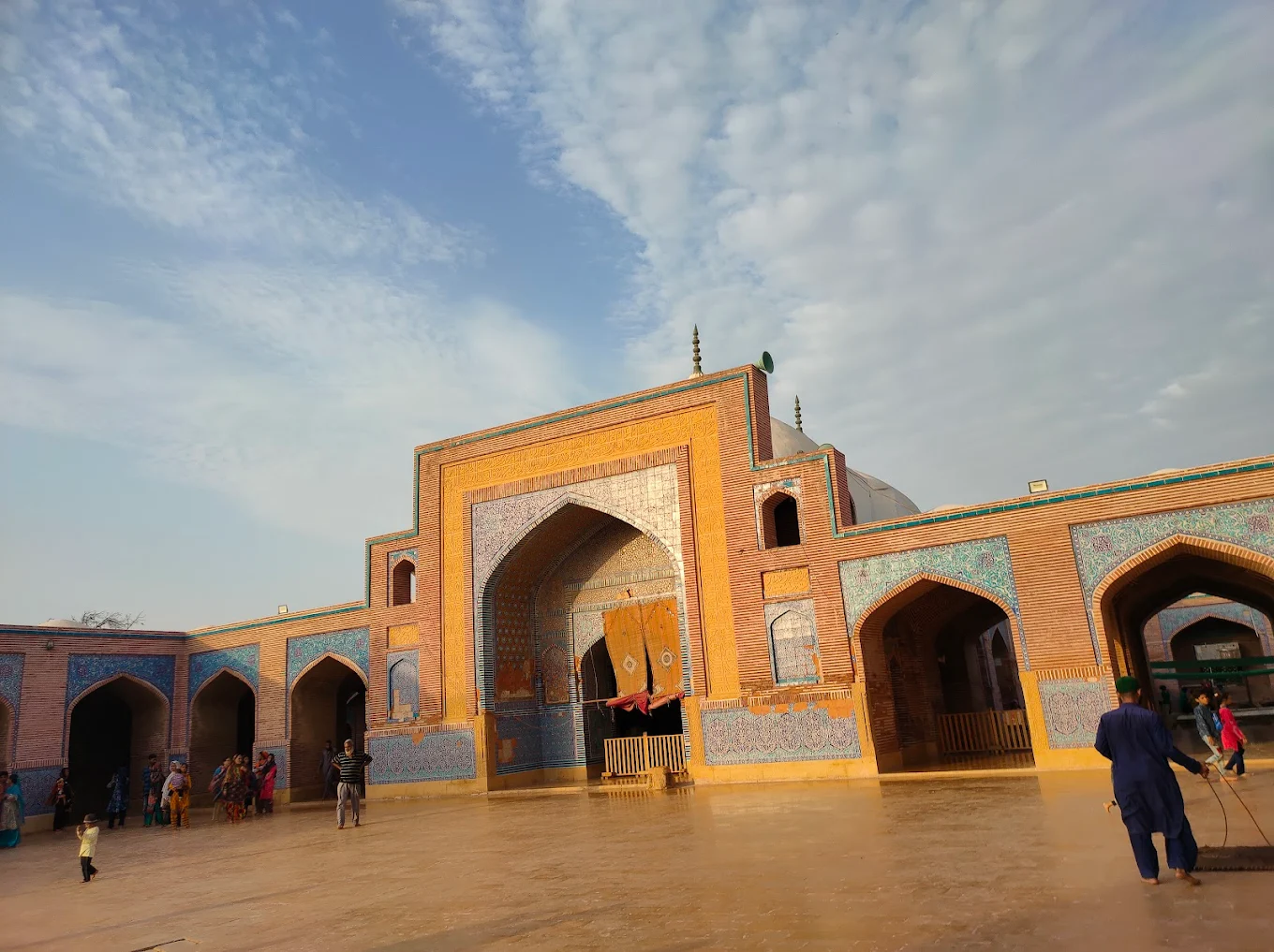 Entrance view of Shah Jahan Mosque in Thatta with detailed blue tile work, brick façade, and people walking on the sunlit courtyard under a partly cloudy sky