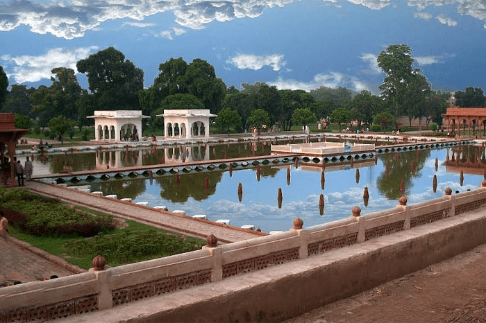 Panoramic view of Shalimar Gardens in Lahore featuring reflective water pools, Mughal-style pavilions, lush greenery, and blue sky with clouds
